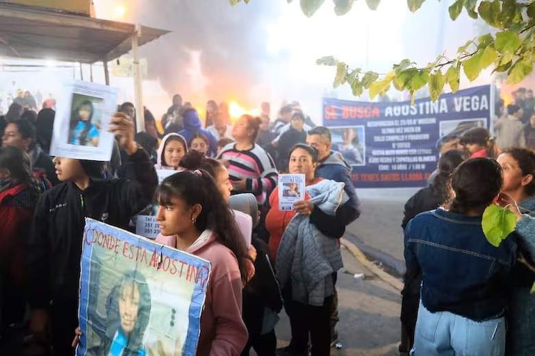Grupo de mujeres sosteniendo carteles que buscan a Agostina Vega, en medio de un ambiente tenso con humo y llamas de fondo.