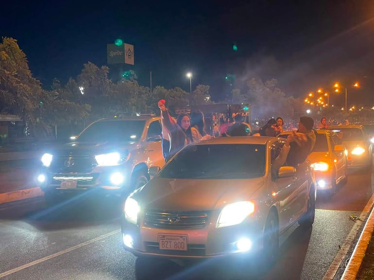 Mujer con bandera en un auto elevado y hombre en el borde, celebrando en una noche festiva llena de luces y humo.