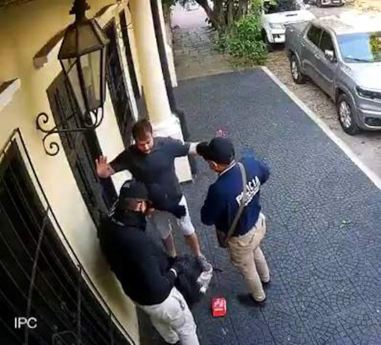 Tres hombres en ropa oscura; uno con camiseta azul de 'Policía' habla a otro que levanta las manos.