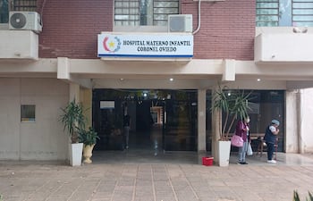 Entrada del Hospital Materno Infantil con dos figuras humanas, una habla y otra limpia con escoba, rodeados de vegetación.