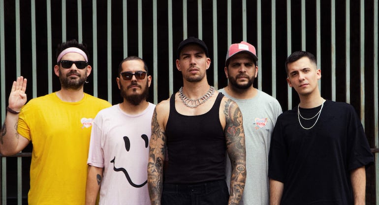 Cinco hombres en camisetas coloridas, entre ellos uno con tatuajes visibles y otro con gafas de sol, posan en un entorno urbano.