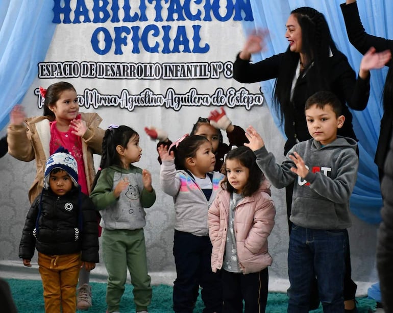 Cinco niños aplauden con sonrisas en un aula decorada, mientras dos adultas dirigen la actividad al fondo.