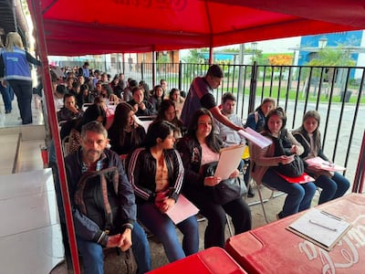 Asistentes sentados bajo un toldo rojo, mujeres y hombres parecen tranquilos, algunos sosteniendo documentos mientras esperan.