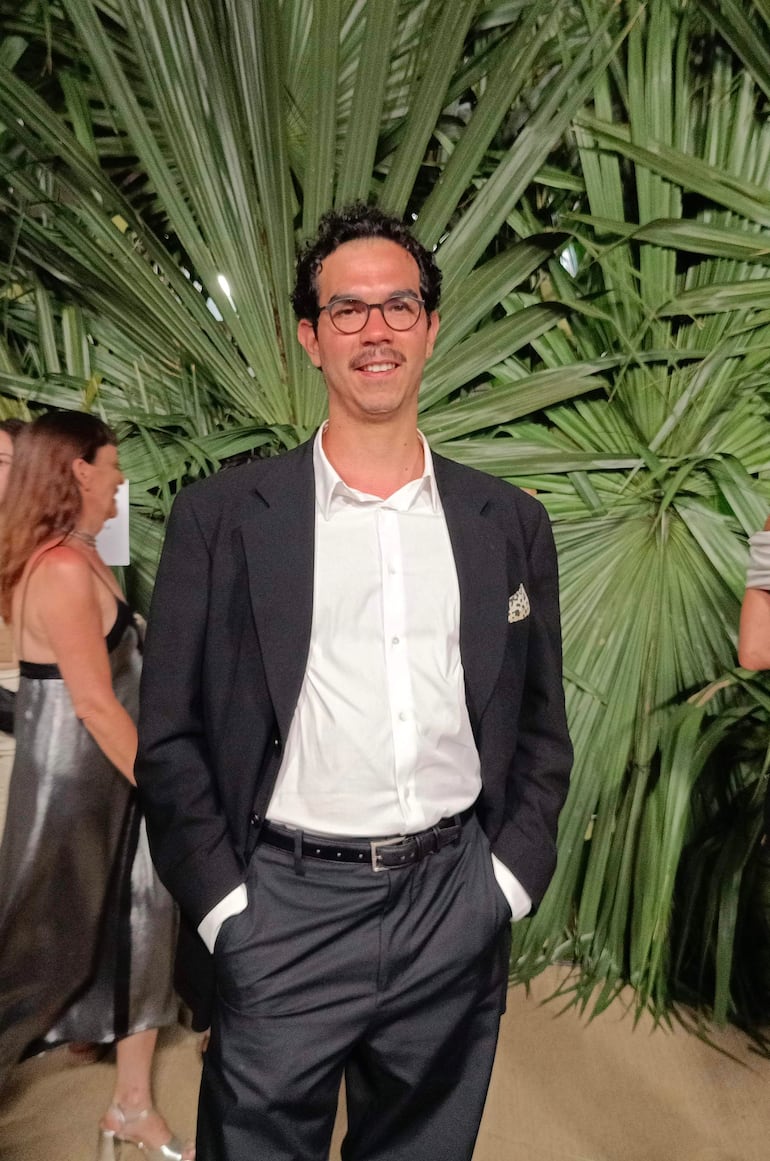Juanjo Pereira en traje oscuro, feliz y con gafas, en un evento social con una mujer de vestido plateado al fondo.