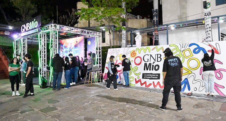Dos personas conversando en primer plano, una con camiseta verde y otra con vestimenta oscura, mientras otros trabajan en un stand iluminado.