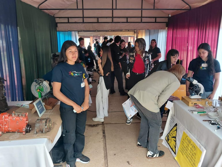 Mujer de pie en camiseta oscura y pantalones negros presenta exhibiciones en un ambiente colorido con jóvenes atentos.