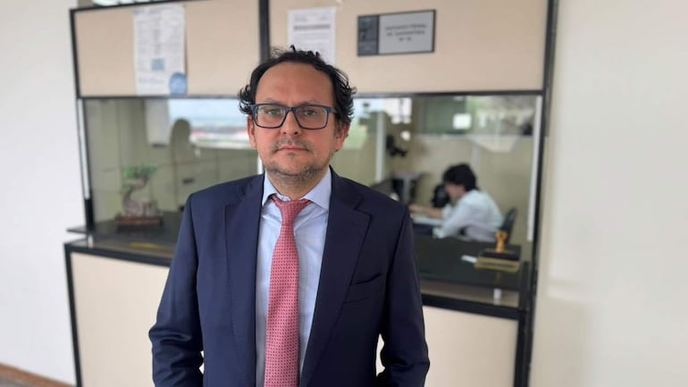 Derlys Martínez, con traje oscuro y corbata rosa, de pie en oficina, mientras un colega trabaja sentado al fondo.