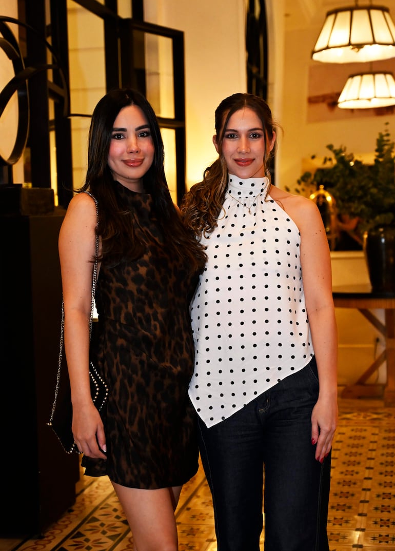 Mujeres sonriendo, una con vestido oscuro y otra con blusa blanca con lunares, en un ambiente elegante y festivo.