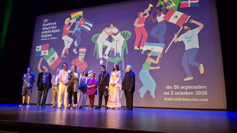 Grupo de ocho personas en el escenario, con Narciso hablando mientras otros escuchan en un ambiente festivo.