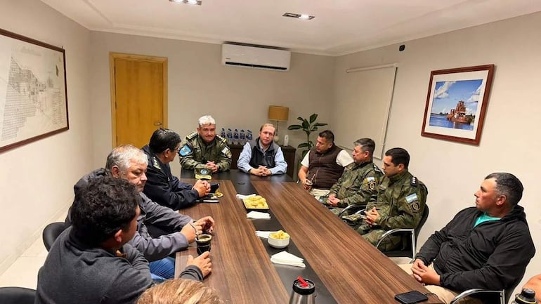 Seis hombres, algunos en uniforme, discuten en una reunión. Un hombre de cabello claro habla mientras otros lo escuchan. Ambiente cálido y mesa con galletas.