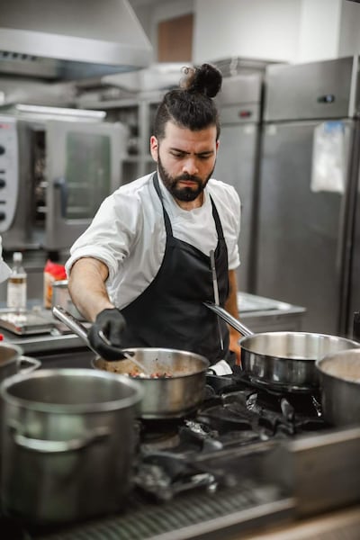 Alejandro Calcaterra, chef con barba y moño, trabaja en cocina industrial con delantal negro y camisa blanca, enfocado en una olla.