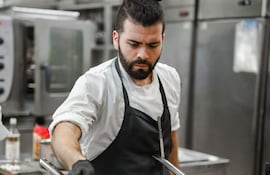 Alejandro Calcaterra, chef con barba y moño, trabaja en cocina industrial con delantal negro y camisa blanca, enfocado en una olla.