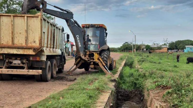 Camión amarillo y excavadora John Deere trabajando en un entorno rural con ganado al fondo y cielo parcialmente nublado.
