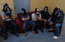 Hombres con capuchas negras que dicen 'Policía Nacional' están sentados en un sofá rojo en una habitación con paredes amarillas.