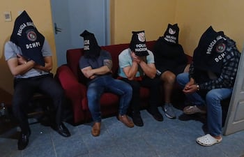 Hombres con capuchas negras que dicen 'Policía Nacional' están sentados en un sofá rojo en una habitación con paredes amarillas.
