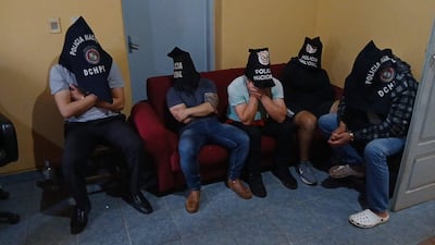 Hombres con capuchas negras que dicen 'Policía Nacional' están sentados en un sofá rojo en una habitación con paredes amarillas.