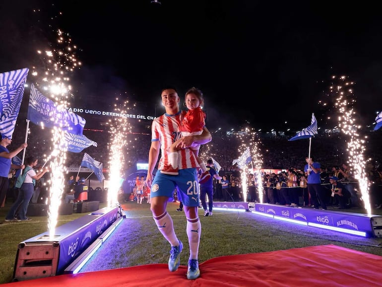 Jugador de fútbol con camiseta roja y blanca sostiene a un niño en un ambiente festivo, rodeado de banderas y luces.