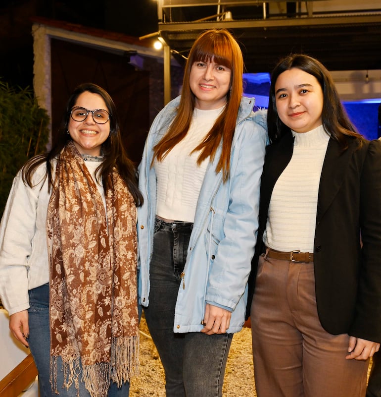 Tres mujeres sonrientes, una con blusa blanca y bufanda, otra en abrigo claro, y la última en blusa blanca con chaqueta negra.