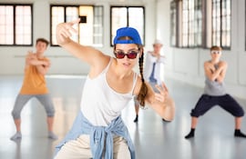 Un grupo de adolescentes baila hip hop en un estudio espacioso. Una chica con una gorra de béisbol azul brillante, gafas de sol y una camisa vaquera atada a la cintura es el centro de atención.