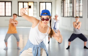 Un grupo de adolescentes baila hip hop en un estudio espacioso. Una chica con una gorra de béisbol azul brillante, gafas de sol y una camisa vaquera atada a la cintura es el centro de atención.