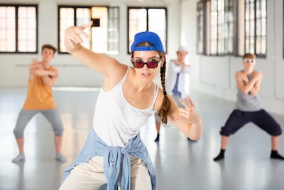 Un grupo de adolescentes baila hip hop en un estudio espacioso. Una chica con una gorra de béisbol azul brillante, gafas de sol y una camisa vaquera atada a la cintura es el centro de atención.