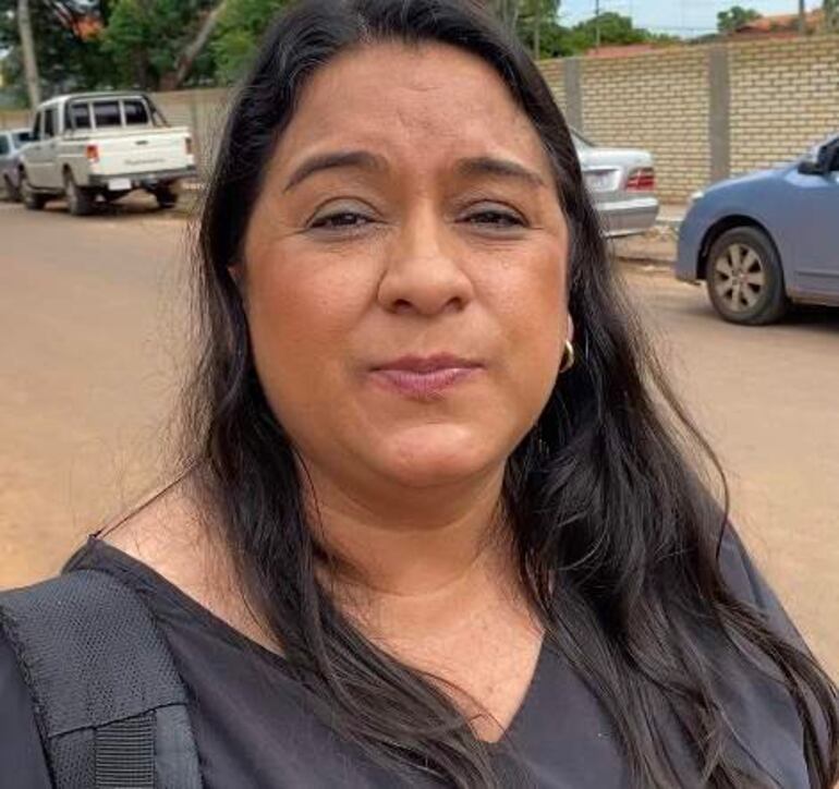 Mujer de cabello largo y oscuro, vestida con top negro, en calle con vehículos estacionados y ambiente cotidiano.