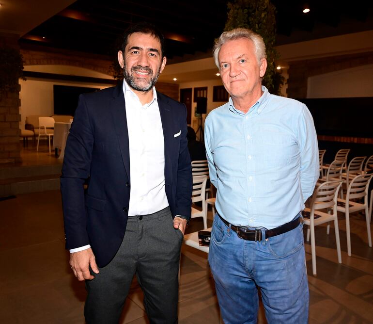Hombre con barba en chaqueta oscura sonríe junto a otro hombre canoso con camisa azul en un ambiente iluminado e informal.