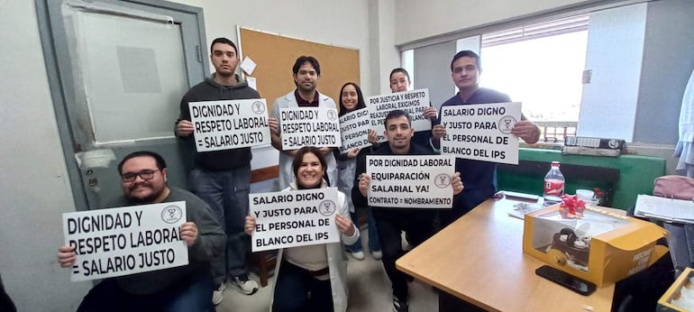 Grupo de profesionales del IPS en reunión, sosteniendo carteles de protesta por dignidad y salario justo, expresiones de seriedad.