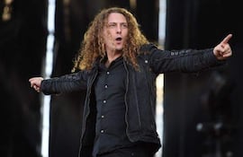 Fabio Lione con cabello rizado y largo, camisa negra y chaqueta, interactúa con la audiencia durante su actuación en un ambiente de concierto.