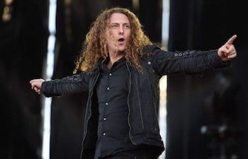 Fabio Lione con cabello rizado y largo, camisa negra y chaqueta, interactúa con la audiencia durante su actuación en un ambiente de concierto.