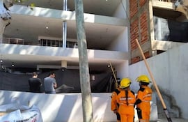 Tres hombres en obra de construcción; dos bomberos con cascos amarillos y ropa naranja, mirando la estructura en construcción.