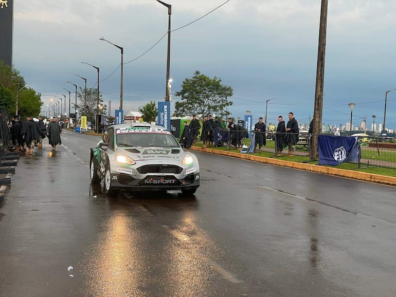 Automóvil de carrera M-Sport avanza mientras asistentes con capas observan bajo lluvia y pavimento mojado.