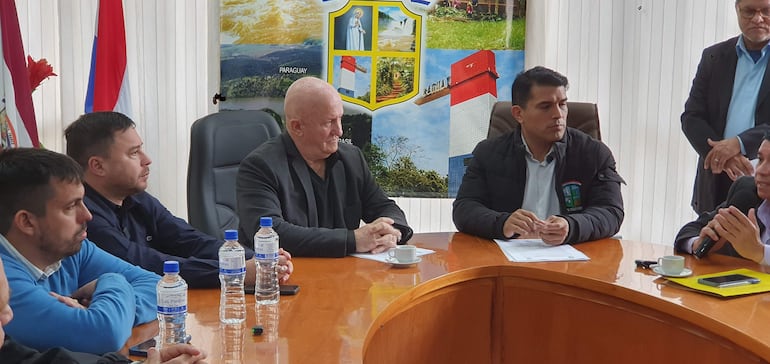 Hombres sentados en mesa discutiendo, uno de cabello canoso vestido de negro, banderas de Paraguay al fondo.