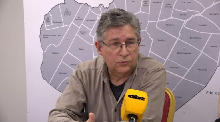 Hombre canoso con gafas sostiene micrófono amarillo en mesa, mientras mapa de Asunción está visible detrás.