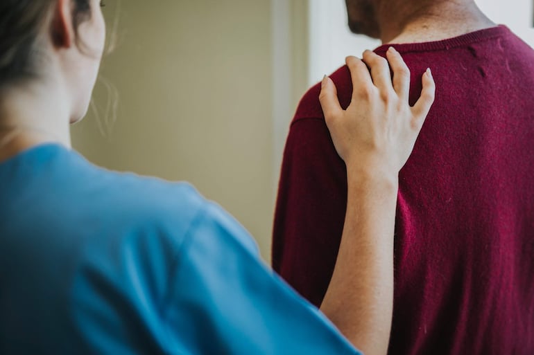 Persona de camiseta burdeos y mujer de camiseta azul conversan, ella apoya su mano en su hombro en un ambiente interior.