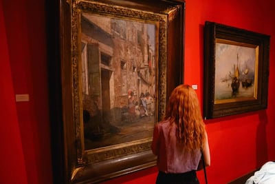 Mujer de cabello rizado y rojo, vestida con blusa morada, observa un cuadro en una galería con fondo rojo.