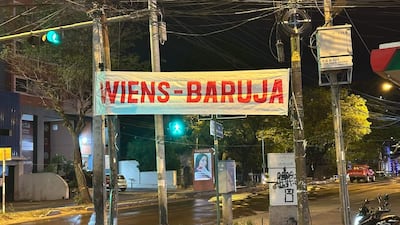 Banner iluminado con inscripción 'WIENS-BARUJA' en la calle, rodeado de árboles y señalización urbana.