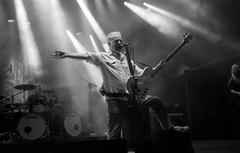 Músico de Carcass en el centro del escenario, sosteniendo un bajo y extendiendo el brazo hacia el público. Luces brillantes y humo de fondo.