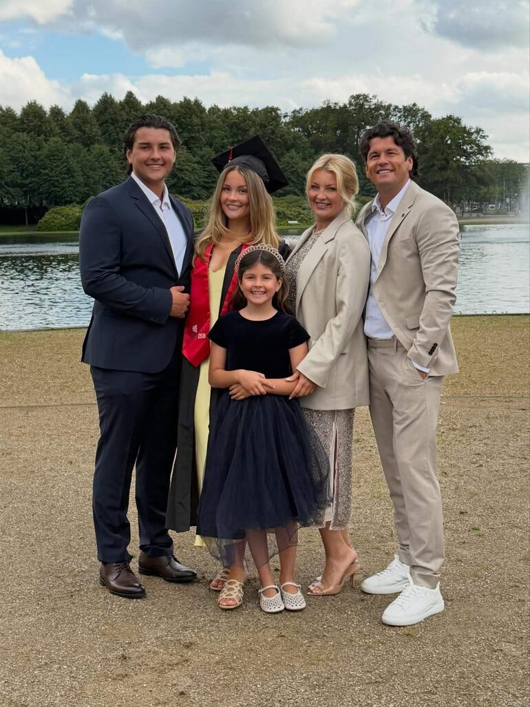 Isabel en toga negra y birrete, sonríe con diploma, rodeada de familiares felices en un parque con lago y árboles.