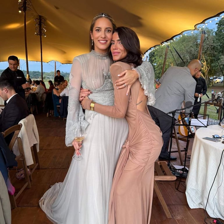 Dos mujeres sonrientes, una con vestido blanco y otra en beige, abrazadas con copas de vino en un evento al aire libre.
