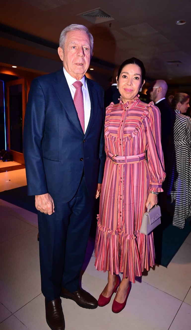 Pareja sonriente en evento elegante; hombre con traje oscuro y corbata rosa, mujer con vestido de rayas rosa y blanco y bolso pequeño.