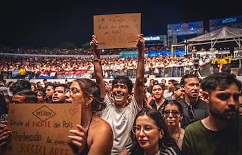 La imagen muestra a un grupo de personas en un evento, posiblemente un concierto, con una gran cantidad de público visible al fondo. Hay al menos tres individuos destacables en primer plano:
1. **Joven con cartel**: A la izquierda, un joven sostiene un cartel con una pregunta relacionada con la compra de un auto, mostrando una expresión de alegría. Este parece ser el foco principal de la imagen.
2. **Chica a la izquierda del joven**: Ella sostiene otro cartel que dice "Desde el NORTE del País, Viviendo la experiencia del RECICLARTE", con una expresión de entusiasmo.
3. **Hombres y mujeres en segundo plano**: Varias personas están más atrás; la mayoría presenta expresiones emocionadas, mirando hacia el escenario.
El ambiente es exterior, con iluminación tenue y una multitud animada. Hay banderas visibles que indican la posible presencia de hinchas, pero no se identifican equipos de fútbol claros. No hay monumentos ni estadios reconocibles en la imagen.