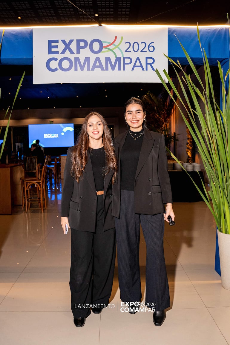 Dos mujeres sonrientes con blazers negros posan juntas, rodeadas de un ambiente festivo y decoración del evento Expo Comampar 2026.