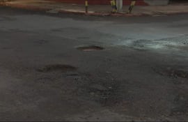Calle con baches visibles en el asfalto, sin presencia de personas, con ambiente oscuro y acera al lado.