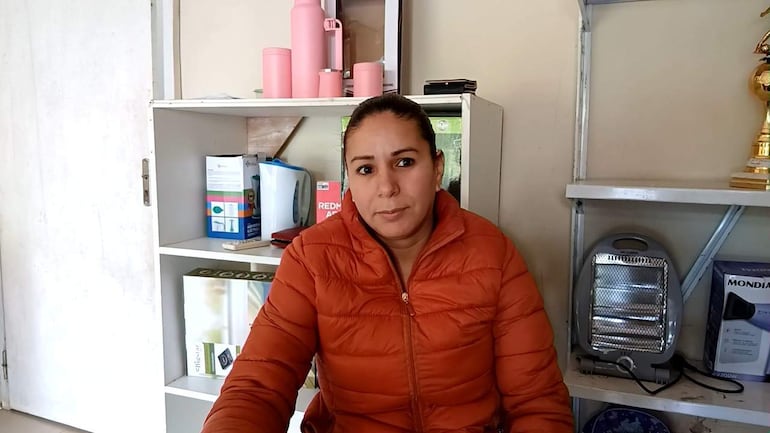 Dahiana Bogado, con chaqueta acolchada naranja, sentada en una oficina, mirada seria, en un entorno sencillo con estantes de utensilios.