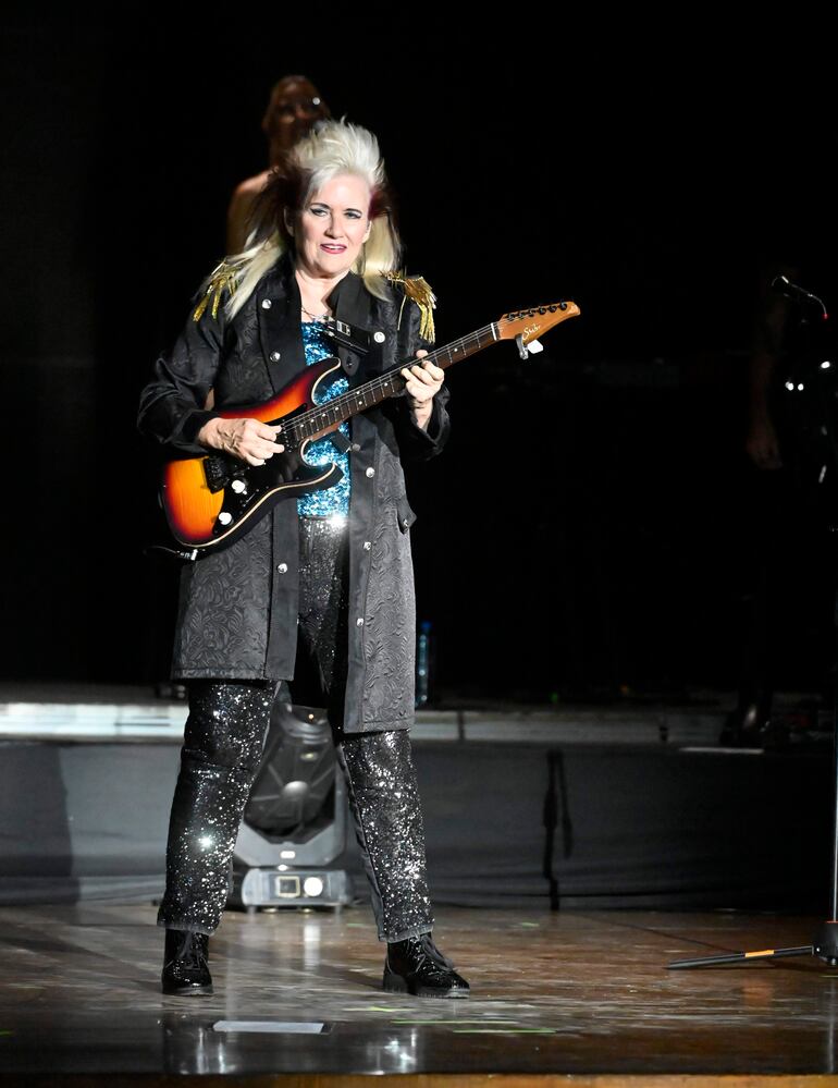 Mujer rubia en atuendo negro brillante sostiene guitarra eléctrica, destacando en concierto con luces de escenario.