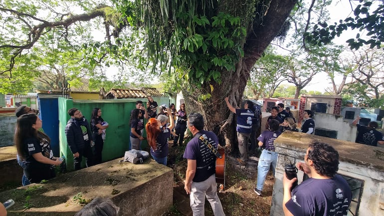Hombres y mujeres con camisetas oscuras escuchan atentos a un hombre de perfil frente a un mausoleo, bajo un árbol frondoso.