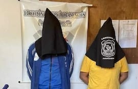 Dos hombres con sacos negros cubriendo sus cabezas, uno en camiseta azul y otro en amarilla, frente a un panel policial.