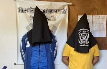 Dos hombres con sacos negros cubriendo sus cabezas, uno en camiseta azul y otro en amarilla, frente a un panel policial.