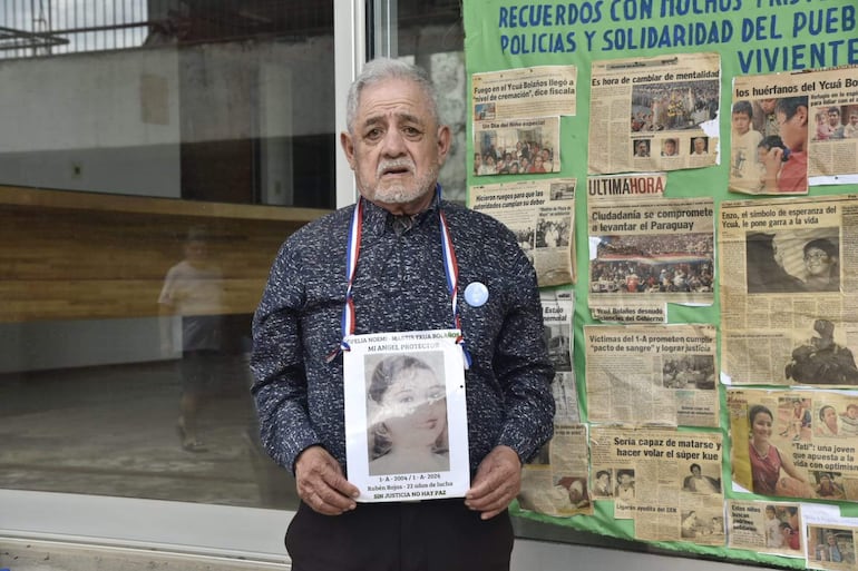 Hombre mayor con camisa azul y medalla sostiene cartel con homenaje, frente a mural de recortes sobre justicia y memoria histórica.